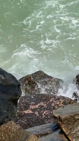 A vertical shot captures the dynamic interaction between the sea and a rocky shoreline in Walgama, Sri Lanka. The scene is dominated by light green-blue ocean water, churning with white foam as waves crash and recede against dark, rugged rocks. The waves periodically engulf parts of the rocks, creating splashes and foamy eddies. Some rocks are partially covered in dark patches, possibly algae or wet areas. A small piece of debris or stick floats momentarily in the water. The video shows no people and appears to be filmed on a bright, clear day. No visible text is present.