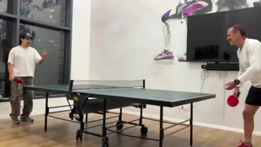 In a brightly lit room, two men engage in a game of table tennis on a green and black table. The man on the left, wearing a white t-shirt, camouflage pants, and a baseball cap, is in the act of hitting the ping pong ball. The man on the right, dressed in a white long-sleeved shirt and dark shorts, prepares to receive the shot. A large window is behind the man on the left, reflecting the interior of the room. On the wall behind the ping pong table and the man on the right, there is a gray artistic mural of an athlete, and a black television is mounted above a sound bar. The floor is light-colored wood.