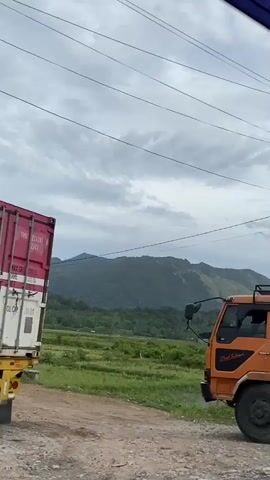 The video depicts an orange truck parked on a dirt road with a background of green fields and mountains under a cloudy sky. The camera pans along the side of the truck, revealing its cargo bed covered with a black tarp. The truck's license plate, BC 8992 EZ, is briefly visible. The camera then continues panning to the landscape, showing power lines, vegetation, and the distant mountain range under the overcast sky.