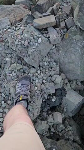 A first-person perspective video shows a person's leg and foot, wearing a grey and yellow hiking shoe, standing on a pile of rocks and rubble. The camera pans up to reveal a body of water in the background, with a sailboat visible in the distance under a cloudy sky. The scene appears to be a rocky shoreline, possibly at a harbor or coastal area. The lighting suggests it may be dusk or dawn.