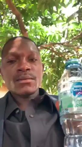 A man holding a bottle of Pure Life water takes a large drink. He is wearing a dark suit jacket and a dark shirt with the top button unbuttoned. The camera is at eye level and close to the man’s face. He is standing under a tree with green leaves.