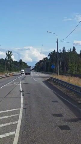 This is a first-person perspective video taken from a vehicle driving on a highway. The highway appears to be in a rural or suburban area with trees lining the right side of the road. The sky is blue with some white clouds. There are other vehicles on the road, including trucks and cars. The road has multiple lanes and is well-maintained, with clear lane markings. There are streetlights visible along the side of the road. A blue traffic sign with a white arrow pointing up is visible on the right side of the road. The video captures the continuous motion of the vehicle as it travels forward.
