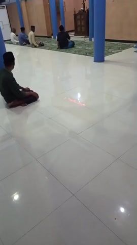 A group of men are sitting on the floor in what appears to be a mosque. The floor is tiled and shiny. There are blue pillars visible, as well as what looks like a mihrab with Arabic calligraphy above it and an LED display that reads "Assalatu'l Mustaqim." One man in the foreground is wearing a green shirt and a black cap and is facing away from the camera, with his hands clasped. Other men are seated further back in the room, facing the mihrab. The lighting is dim.