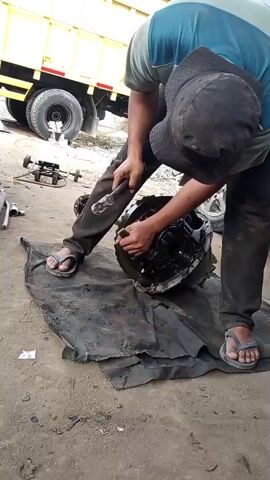 A person wearing a blue and grey shirt, dark pants, a black hat, and sandals is working on a large, black, oily mechanical component that is resting on a black tarp on the ground. The person is holding a metal object, possibly a hammer, and appears to be striking the component. Another person can be seen in the background. A yellow truck is parked behind the person, and there are other tools and mechanical parts scattered around the scene. The ground is dirt.