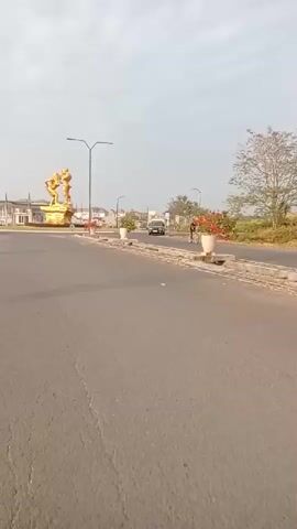 The video shows a road with a roundabout in the background. There is a large gold statue of two figures in the center of the roundabout. Several cars and a motorcycle are visible on the road and in the roundabout. The sky is overcast. The overall scene appears to be a suburban or semi-rural area.