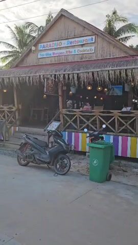 The video shows a side view of a restaurant called "Paradiso Restaurant" which appears to be built in a rustic, tropical style with a thatched roof. In front of the restaurant, a black scooter is parked. To the right of the scooter, a green trash bin with a white symbol is visible. The restaurant has a wooden porch with colorful striped railings. There are lights hanging under the porch roof. Palm trees are visible behind the restaurant, and the sky is partly cloudy.