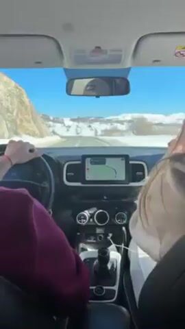 Inside a car driving on a snowy road on a sunny day, captured from the backseat. A person wearing a purple shirt is driving the car, visible holding the steering wheel and operating the gear shift. A passenger wearing a light brown hoodie is seated in the front passenger seat, their back visible. The car's dashboard displays a navigation system with a visible route. Through the windshield, the road ahead is bordered by snow-covered hills under a bright blue sky. A road sign warning of deer crossing is visible on the right. The interior of the car is clean and modern. Upbeat music plays in the background.