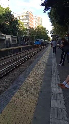 The video shows a train station platform with people waiting as a blue and silver train approaches. The platform has a textured yellow safety strip, and there are train tracks and buildings visible in the background. A man walks on the platform, and two women are leaning on a pole. There are trees on both sides of the tracks. The sky is blue with some clouds.
