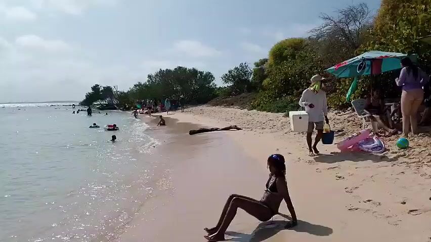 The video shows a crowded beach on a sunny day. The beach has white sand and clear water with people swimming and playing in the water. There are people sitting on the beach under umbrellas. A woman in a bikini is sitting on the beach in the foreground. Two children are running along the beach near the shore.