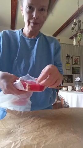 A woman in a light blue sweatshirt is shown from the chest up, preparing raw tuna steaks. She is standing in what appears to be a kitchen or dining area with exposed wooden beams on the ceiling. To her right, a white tablecloth covers a table with cups and decorative items. The woman is holding a white package in her hands, from which she is pulling out bright pink tuna steaks. One steak has already been placed on a sheet of brown parchment paper on a wooden surface in front of her. The lighting suggests it is daytime, and the indoor setting implies a home environment. The woman's expression is neutral, focused on the task at hand. No visible text is present in the media.
