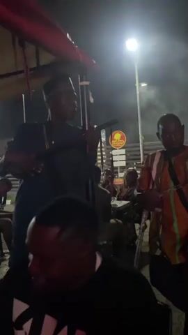 The video shows a nighttime scene outdoors, likely a street or market area, illuminated by artificial lights and with some smoke present. In the foreground, partially visible, is a person wearing a black shirt. Behind them, a man in a dark garment and a traditional cap holds a long, dark object, possibly a musical instrument or a staff. To the right, another man, wearing a brightly colored striped shirt with orange and green, uses crutches and also holds a long, dark object. In the background, a food stall with the sign "SPECIAL SUYA" is visible. Other people are present in the background, some seated at tables. The overall atmosphere appears to be lively, possibly a performance or social gathering.