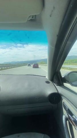 The video shows a view from inside a car traveling on a highway. The sky is blue with scattered clouds. In front, two cars are visible driving in the same direction. The dashboard of the car taking the video is grey and shows some signs of wear. The side mirror shows the red car next to it. In the background, mountains and greenery are visible on either side of the road.