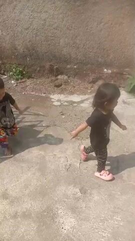 Two young children are playing outside on a sunny day. The first child, a boy wearing a black t-shirt and patterned shorts, runs across a concrete surface. He is followed by a girl in a black t-shirt and striped leggings, wearing pink sandals. The children are in an outdoor area with a textured wall and some vegetation in the background. The ground is a mix of concrete and gravel. The boy then walks onto a tiled walkway beside a red-walled building.