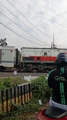 A group of motorcyclists, many wearing helmets and some with Grab uniforms, are stopped at a railroad crossing. A train is passing by. The sky is cloudy. The scene appears to be a street in a populated area, possibly in Southeast Asia, given the presence of Grab. One motorcyclist in the foreground is wearing a black jacket with green accents and the Grab logo. The train cars are light gray and white with orange trim.