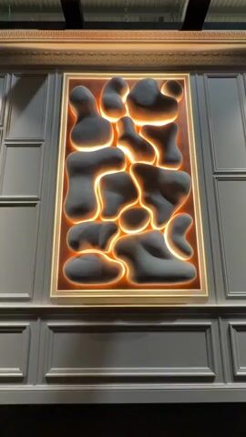 The video shows a piece of wall art. The art is set in a rectangular gold frame, and it is lit from behind with a warm orange light. Inside the frame are several irregularly shaped, matte gray objects. They appear to be three-dimensional, as if they are raised from the background. The wall behind the art is paneled in a gray color with a rectangular pattern. The camera slowly zooms in to get a closer view of the art. The lighting is slightly dim, giving the scene a soft glow.