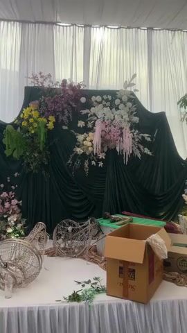The video shows a wedding or event setup in progress. The scene is an indoor venue, possibly a large tent, with white draped fabric forming the ceiling and walls. The primary focus is a stage area decorated with lush floral arrangements in shades of white, pink, and green. There are also decorative trees with fairy lights, and ornate furniture. In the foreground, there are boxes, ladders, and other setup materials, indicating that the event is not yet complete. A person is seen working on the decorations, suggesting that staff are actively preparing the space. The lighting appears to be a mix of ambient artificial light and decorative lights. The overall atmosphere is one of anticipation and preparation for a celebratory event.