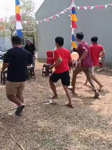 A group of people are playing a game outdoors. They are holding balloons and seem to be running and passing them to each other. There are several chairs set up, and decorative flags are strung across the area. The ground is covered in gravel and dry leaves. A building is visible in the background.