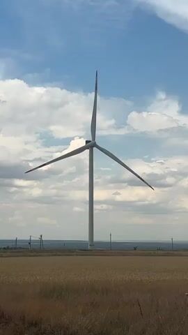 A vertical video shows a single white wind turbine with three blades in a field of dry grass. The sky is blue with white clouds. In the background, there are power lines and some distant trees. There is a road with a dark-colored car driving on it, and it appears to be a rural setting. The wind turbine is stationary. The video appears to have been filmed on a sunny day.
