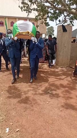The image captures a funeral procession in Ladura Foron, Nigeria, during the daytime under sunny weather. The central focus is on four men, dressed in dark blue suits, white shirts, and ties, with white gloves, acting as pallbearers. They are carefully carrying a wooden coffin, which is draped with a green and white flag, emblematic of the Nigerian national flag. One of the pallbearers in the foreground is wearing a white face mask.

The men are walking on a reddish-brown dirt path, which appears to be unpaved. In the background, numerous individuals are gathered, some observing the procession, others moving alongside. Their attire varies, including casual wear and patterned traditional garments. To the left, a building with a distinctive red roof and light-colored walls is visible, possibly a religious structure. Large trees flank the scene, providing some shade. The overall atmosphere suggests a solemn community event, as implied by the nature of a funeral. A motorbike or moped is partially visible among the crowd on the right side of the frame. No discernible text is visible in the image.