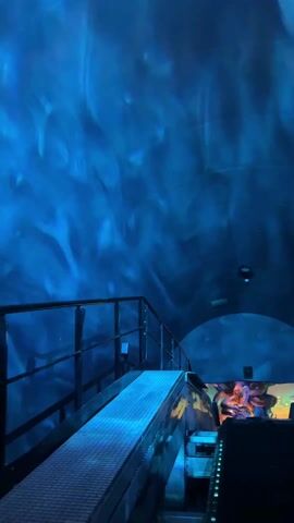 The video shows a view from a moving walkway in an indoor location with blue lighting effects on the ceiling, resembling water. A metal railing is visible on the left. In the distance, people are seen on a ride or attraction, with colorful elements suggesting a theme park or entertainment venue. The focus is on the lighting and movement of the walkway.