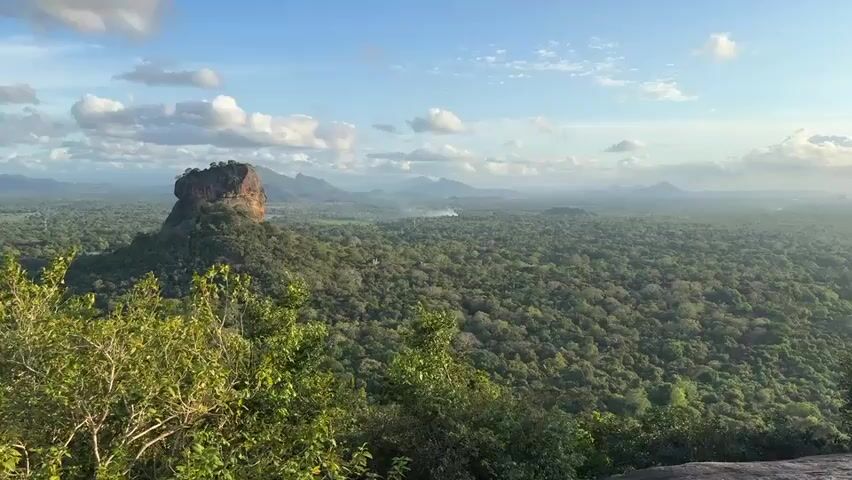 A wide shot shows a large rocky hill surrounded by a dense forest. In the background, there are mountains on the horizon under a sky with scattered clouds. The foreground shows lush green trees.  People can be heard speaking in the background.