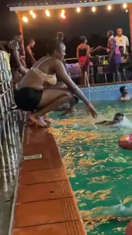A group of people are gathered around a swimming pool at night. Several people are swimming in the pool, some using inflatable inner tubes. A woman wearing a light-colored bikini top and dark shorts is crouching at the edge of the pool. She is reaching out toward the water with one hand. Other people are standing or sitting near the pool, some engaging in conversation. String lights hang above the pool area, illuminating the scene.