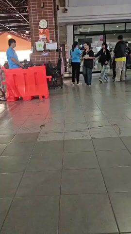 The video shows a man in a blue shirt and gray pants walking away from the camera, then turning to face the camera and making a gesture. He is in a tiled walkway. To his left, there is a red plastic barrier. Behind the barrier, a man in a blue shirt is standing and talking to someone off-camera. Several other people are visible in the background, walking in the walkway and near a building entrance. The environment appears to be a public indoor or semi-outdoor space, possibly a market or transit station. The lighting is bright.