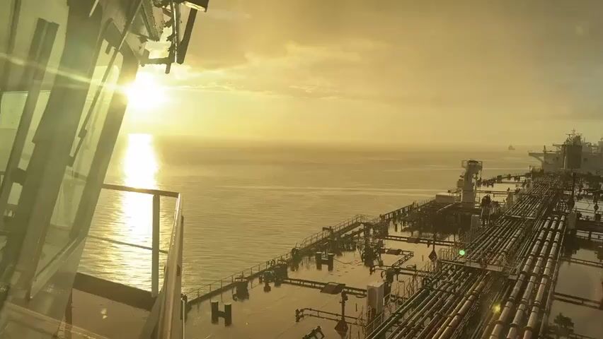 The video opens with a view from a ship's bridge, looking out over the deck towards the sea at sunset. The sun is low on the horizon, casting a golden glow across the water and sky. The deck of the ship is filled with a network of pipes and equipment. In the distance, other ships can be seen on the horizon. The scene is calm and serene.