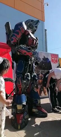 A large Transformers Optimus Prime character costume with red and blue flames is standing outdoors, facing slightly right, with a small girl in a white shirt and a white bow in her hair at its left, interacting with its right arm. The costume has a black upper body, red shoulders and chest with black flame details, and blue accents on the lower body and legs, also with red flames. The girl is holding what appears to be a light saber or similar toy in her left hand, and her right hand is near the costume's right hand. Another person, partially visible, is standing to the right of the costume, bending forward. A red tent or banner is visible behind the costume on the left, and a large white banner with a Transformers logo and characters is behind it on the right. A tall building with an orange upper section and lighter blue lower section is in the background. The sky is clear and blue. Another child in a gray cap is to the left of the costume, holding up a black ball. There are other people in the background, out of focus.