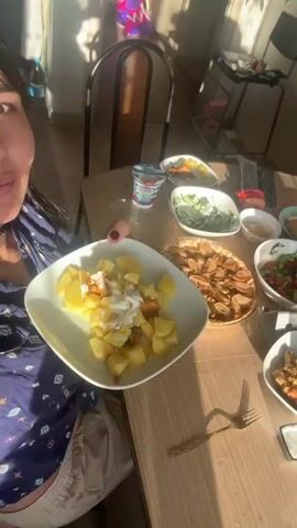 The video shows a table laden with food, including what appears to be an apple pie, a salad, potatoes with a creamy sauce, and a yogurt cup. There are also several plates, bowls, glasses, and a napkin. In the foreground, a person's hand with painted fingernails is visible. The background shows a room with furniture and natural light streaming in. The final shot is a close-up of a young woman's face. The overall scene suggests a meal or gathering.