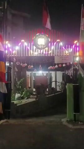 A short video showcases a building entrance at night, adorned with festive lights and flags. The entrance is framed by vertical elements with a wavy string of multi-colored lights draped across the top. A circular emblem with lettering, partially obscured by the lights, is visible in the center. Below this, a row of white circular decorations with red accents hangs across the building's facade. The Indonesian flag, red and white, is prominently displayed on a flagpole above the entrance. Two additional flags, featuring multiple horizontal color bands, are positioned to the left and right of the entrance. The building itself appears to have a traditional architectural style, with a visible doorway at the center. The scene is illuminated by the colorful lights, casting a festive glow on the area. The ground in front of the entrance is paved and appears to be a street or pathway. Vegetation is visible to the left of the entrance. The overall impression is one of celebration or a special occasion.