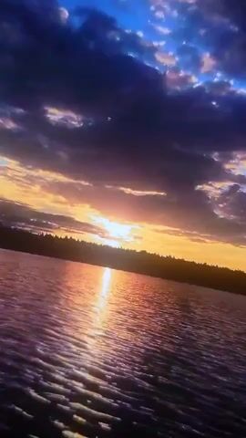 The video is a first-person perspective of a sunset over a lake. The sun is low on the horizon, casting a golden glow across the water. The sky is filled with dramatic clouds, ranging from deep purple to bright orange. A person's shoulder and hood are visible in the lower right corner of the frame, suggesting they are observing the sunset from a boat or shore. The water is rippling, reflecting the colors of the sky. The shoreline in the distance is silhouetted by the sunset.