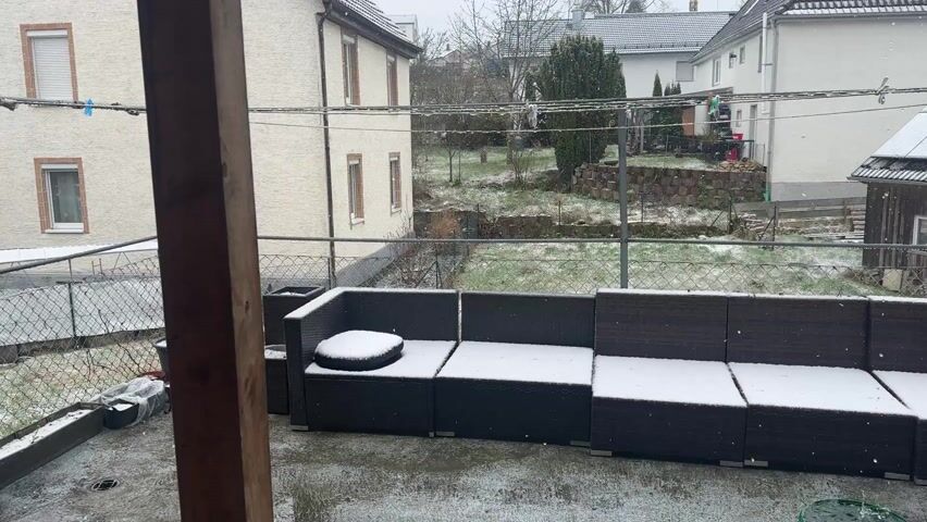 The camera is positioned inside a covered porch, looking out onto a snowy backyard. The porch has a dark-colored wicker patio set covered in a thin layer of snow. A wooden post is partially visible on the left side of the frame. Beyond the porch, the backyard is a mix of grass and snow. A chain-link fence separates the yard from the neighboring properties, which include houses with light-colored facades and gray tiled roofs. A few trees and bushes are scattered throughout the yards, some also dusted with snow. Snowflakes are visibly falling.