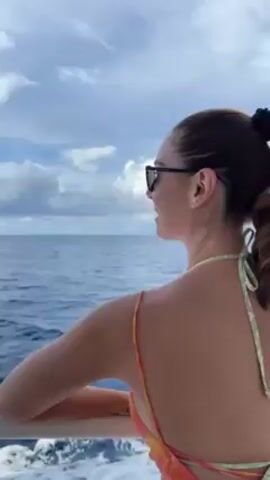 A woman wearing sunglasses and a floral print top looks out at the ocean from a boat. She has her hair in a ponytail. In the distance, a large cruise ship is visible. The boat appears to be moving towards the cruise ship.  A boy wearing a black shirt is visible looking down and to the side.  The sky is cloudy. The name "Celebrity Beyond" is visible on the cruise ship.