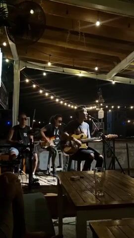 A three-person band is playing live music at an outdoor venue at night. The band consists of two guitarists and a keyboardist, all seated. The lead singer is playing an acoustic guitar and singing into a microphone. String lights are hung above the band, creating a warm ambiance. A table with a glass of water is in the foreground. People can be seen sitting at the table, watching the performance. The overall atmosphere is relaxed and casual.