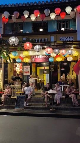 The video shows the exterior of a bar called Iberico in what appears to be Vietnam. The bar is decorated with colorful lanterns hanging above the entrance, and people are sitting at tables outside. Inside, there is a bar area with people sitting and standing. A server is standing in the doorway. A chalkboard menu is placed near the front of the establishment. Other people can be seen walking by on the sidewalk. The sky is a deep twilight blue.