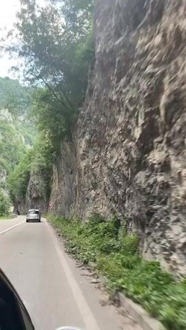 This is a first-person perspective video taken from inside a vehicle traveling on a winding road through a mountainous, forested area. The road is paved and has a white line dividing the lanes. Several cars are visible on the road ahead, suggesting moderate traffic.

The dominant subject is the dramatic natural scenery. A massive, rugged rock face rises vertically to the right of the road, covered in patches of vegetation and showing signs of erosion. To the left, steep, tree-covered mountains rise, creating a canyon-like environment. The trees are lush and green, indicating a temperate climate and likely a time of year with full foliage, possibly spring or summer. The sky is overcast, suggesting a cloudy or possibly rainy day, but the overall light is bright enough for clear visibility.

The activity is vehicular travel, specifically a car journey through a scenic but potentially hazardous route, as indicated by road signs. One sign depicts a sharp curve, and another, a triangular warning sign with "100 m" below it, likely indicates a sharp curve or hazard within 100 meters. The presence of these signs and the narrowness of the road, hugging the cliff face, suggest a potentially challenging drive.

Notable details include the texture of the rock face, the dense green of the foliage, and the reflective surfaces of the cars. The camera perspective, looking out the side window and partially capturing the car's interior elements like a side mirror, adds to the immersive, on-the-go feel of the video. There are no people clearly visible in the frame. The overall mood conveyed is one of awe at the natural landscape and the experience of a road trip through dramatic terrain. The location is identified as Prijepolje Municipality, Serbia.