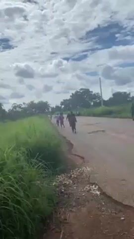 The video shows a group of people walking along a paved road with some potholes. The road is flanked by green vegetation on either side. The sky is cloudy with patches of blue visible. The camera seems to be positioned inside a vehicle, and part of the dashboard is visible in the bottom right corner. The people are scattered along the road, walking in the same direction.