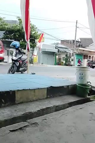A motorcycle with a person wearing a blue helmet is parked on the side of the road. A red truck with a yellow cargo bed is driving away from the camera. In the background, there are several buildings, including a store with a green awning and a white garage door. A white SUV is driving past the motorcycle. There are two buckets next to the road, one white and one green. The scene appears to be a street in a town or city. The video is taken from a low angle, looking up at the street. There are red and white flags hanging from poles on either side of the frame.