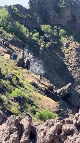 The video shows a view looking down into a volcanic crater. There is steam or gas emanating from a rocky area on the slope of the crater. Some sparse green vegetation is visible on the crater slopes. In the middle ground on the left side of the image, there are two dark rectangular objects that appear to be solar panels. The foreground is composed of rough, reddish-brown volcanic rock. A wooden railing with rope is visible in the bottom of the frame. The sky is clear and blue.