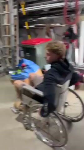 A young man with blond curly hair, wearing a black hoodie, shorts, work boots, and white socks, is sitting in a wheelchair. He turns to his right and then propels the wheelchair forward. The wheelchair is in a cluttered garage with ladders, tools, and various containers. The garage has a concrete floor.