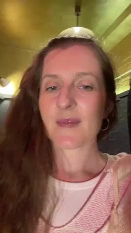 A woman with long, brown hair and wearing a pink sweater over a white shirt is facing the camera. She is looking up and to her left with her mouth open, appearing to be singing or speaking. The background is dimly lit with a golden ceiling and a dark wall. There is a light fixture hanging from the ceiling.