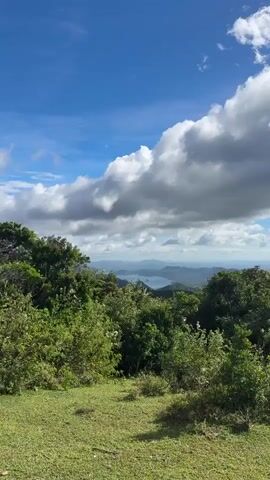 The video presents a scenic view from an elevated position, likely a hilltop or mountain. In the foreground, there's lush green vegetation, including trees and grassy areas. Beyond the vegetation, the landscape opens up to reveal a vista of rolling hills and mountains in the distance. A body of water, possibly a lake or bay, is visible among the hills. The sky is partly cloudy, with a mix of blue sky and large white clouds, some of which are casting shadows on the distant hills. A bird flies by in the middle of the frame.