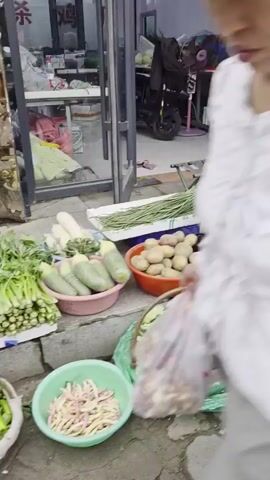 A street market scene unfolds. In the foreground, various vegetables are displayed on the ground and in baskets. To the left, a green plastic bag filled with sliced green beans sits near a basket of potatoes in an orange bowl. Next to the potatoes, a white bowl holds sliced eggplants. Another bowl of sliced eggplants is further to the right. Behind these, a long bunch of green vegetables, possibly green beans or asparagus, is laid out. Further back, a stall has a black electric scooter with a fan behind it. A woman in a patterned shirt and colorful pants stands by a doorway, her hand resting on the frame. Another woman in a red dress bends down to sort tomatoes. In the midground, a white crate holds cucumbers, and a blue bowl contains small white florets, possibly cauliflower. A red basket holds green vegetables, possibly okra. A woman in a yellow plaid shirt and light blue pants walks away from the camera. Another woman wearing a red helmet, a white lace top, and black pants walks past. A woman in a blue denim jacket and white pants is walking towards the camera. She is carrying a brown bag. To her right, a pile of lettuce is displayed on a white surface. In the background, several vehicles are parked, including a blue van. The ground is paved with interlocking bricks.