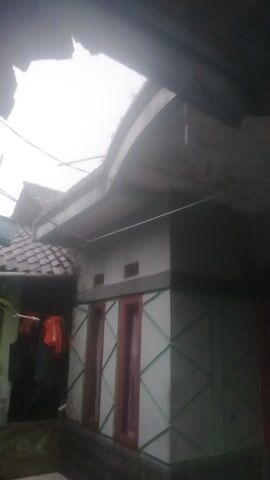 The video features a slow, upward pan of an outdoor or semi-outdoor space, likely an alleyway or a small courtyard situated between residential structures, in Kecamatan Pangalengan, Indonesia. The camera begins by showing an obscured view of the sky and a roof structure, then slowly tilts downwards to reveal more of the architectural elements and foliage.

The lighting is dim and diffused, suggesting an overcast day or perhaps early morning/late afternoon. The sky, visible at the top and left, appears bright but without direct sunlight.

The scene is dominated by building structures. On the right, an overhead structure with a geometric pattern, possibly a metal frame with translucent panels forming a roof or awning, is prominent. Below this, there are sections of walls, some featuring rectangular, possibly wooden-framed, openings or decorative elements. A long, thin white wire or string stretches diagonally across the frame.

Further down and towards the left, a large, vibrant green plant with broad leaves is visible. A striking feature of this plant is a long, drooping, reddish-pink flower stalk, possibly from a species of orchid cactus (Epiphyllum) or similar plant. The plant appears to be growing adjacent to a wall constructed from white concrete blocks. In the darker areas beneath the overhead structures, glimpses of what might be hanging fabric or clothing items in red and green hues can be seen.

No individuals are present in the video, and there is no specific activity taking place. The overall impression is that of a quiet, mundane exterior setting, captured on a cloudy day. There is no visible text.