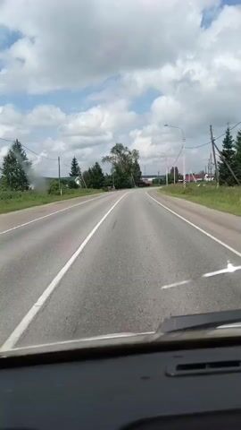 The video shows a POV shot from a vehicle driving on a paved road. The road has two lanes with a solid white line marking the center. The road curves to the right. On the left side of the road, there are trees, a few houses, and some trash bins. On the right side, there are more trees, houses, utility poles with wires, and a lamppost. The sky is partly cloudy with white cumulus clouds against a blue sky. The sun is shining, casting shadows. The dashboard of the vehicle is visible at the bottom of the frame. The scene appears to be a rural or suburban area during the daytime.