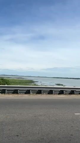 The video pans across a wide body of water with some vegetation along the edge. In the foreground, there is a gray asphalt road and a silver metal guardrail. The sky is partly cloudy with a blue hue. In the distance, rolling hills or mountains are visible. There appear to be some birds on the water. The overall scene suggests an outdoor landscape, possibly a lake or reservoir, viewed from a road.
