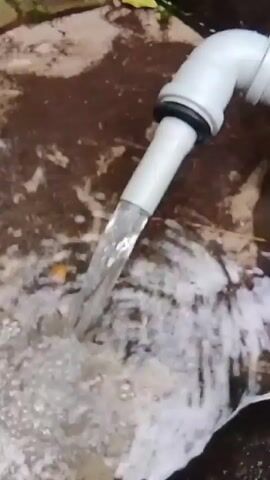 A white hose is spraying water onto a brown, uneven surface, creating a foamy white splash. The camera then pans to the right, showing a gray metal bucket hanging from a wooden structure. Further to the right, the scene reveals a dirt path surrounded by trees and green foliage. The video appears to be shot outdoors, possibly in a forest or garden area.