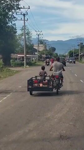 A man and a child are riding on a motorcycle with a sidecar. The man is driving the motorcycle, and the child is sitting in the sidecar. The motorcycle is on a paved road, and there are buildings and trees on either side of the road. In the background, there are mountains. The sky is blue with some clouds. The video is shot from behind the motorcycle, and the camera is moving.