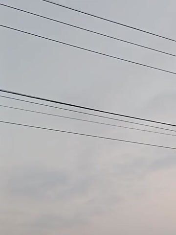 The video shows an outdoor scene dominated by power lines crisscrossing against a cloudy sky. In the lower portion of the video, a building with a corrugated metal roof is visible, along with a glimpse of a storefront with signage that is mostly illegible due to the video's quality and distance. The perspective suggests the video is being filmed from a low angle, looking upwards. There are no discernible people or active interactions depicted.